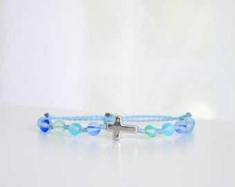May include: A light blue beaded bracelet featuring a silver cross charm. The bracelet has a series of blue and green iridescent beads strung on a light blue cord. The cross is centered on the bracelet.
