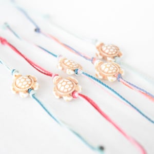 May include: A set of six colourful friendship bracelets, each featuring a small white turtle charm. The bracelets are made with thin string and are tied in a knot at the end.
