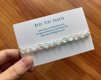 May include: A white iridescent beaded bracelet on a white card with the text "BLUE TIDE CRAFTS" and contact information for the shop. www.etsy.com/bluetidecrafts Instagram/Pinterest: BlueTideCrafts BlueTideCrafts@gmail.com