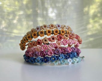 May include: A stack of handmade bracelets, each adorned with small, daisy-like flowers. The bracelets feature a variety of colors, including shades of pink, blue, orange, and white. The bracelets are likely made of a woven or macrame material.
