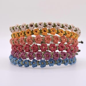 May include: Six handmade bracelets stacked together, each with a floral pattern. The bracelets are in various colours: beige, yellow, orange, red, purple, and blue. They are made of woven material and are likely adjustable to fit different wrist sizes.