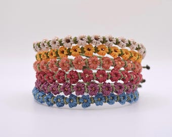 May include: A stack of six handmade bracelets, each featuring a floral design. The bracelets are in a variety of colors, including beige, yellow, orange, red, purple, and blue. The bracelets are made of a woven material and are likely adjustable.