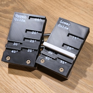 Op de afbeelding: Twee zwarte hoekgeleiders met witte tekst en gradenmarkeringen. Eén geleider is gemarkeerd als "Bovenste geleider" en de andere als "Onderste geleider". De geleiders hebben hoeken van 15 tot 40 graden. Op een houten oppervlak.