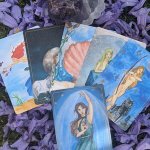 Puede incluir: Un juego de cartas de tarot con ilustraciones coloridas y detalles dorados. Las cartas est&aacute;n dispuestas sobre un lecho de flores moradas. Una carta muestra a una mujer con un vestido verde con la palabra "Protecci&oacute;n" escrita debajo de ella.