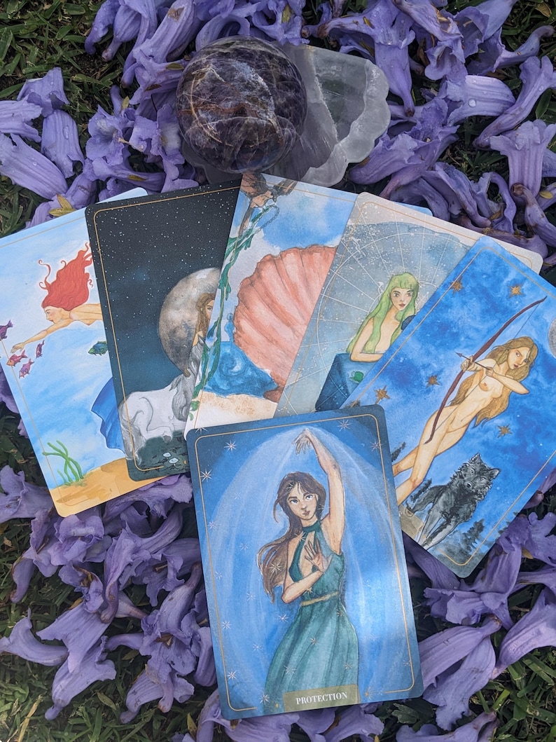 Puede incluir: Un juego de cartas de tarot con ilustraciones coloridas y detalles dorados. Las cartas est&aacute;n dispuestas sobre un lecho de flores moradas. Una carta muestra a una mujer con un vestido verde con la palabra "Protecci&oacute;n" escrita debajo de ella.