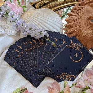 Puede incluir: Una baraja de cartas de tarot con fondo negro y detalles dorados. Las cartas est&aacute;n desplegadas en semic&iacute;rculo, revelando una carta con una luna creciente y la letra "N" en dorado.