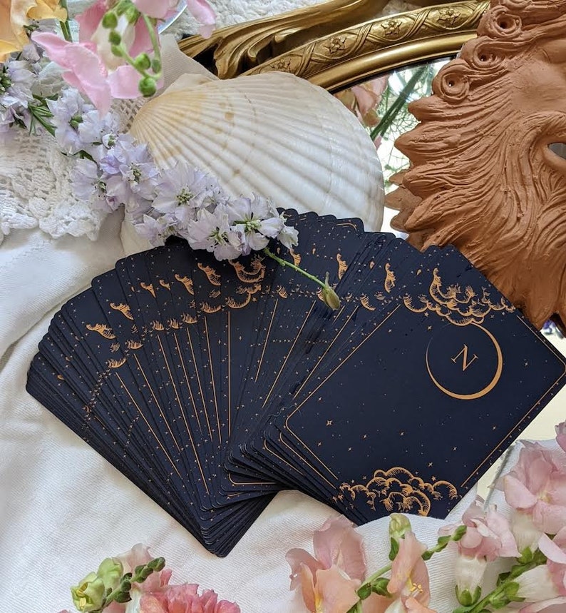 Puede incluir: Una baraja de cartas de tarot con fondo negro y detalles dorados. Las cartas est&aacute;n desplegadas en semic&iacute;rculo, revelando una carta con una luna creciente y la letra "N" en dorado.