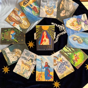 Puede incluir: Un despliegue de cartas de or&aacute;culo con un esquema de color azul, rojo y dorado. Las cartas presentan ilustraciones de mujeres, animales y escenas de la naturaleza. Las cartas est&aacute;n dispuestas en c&iacute;rculo sobre una superficie de terciopelo azul con estrellas doradas. La carta del centro dice "Nymph Oracle Cards".