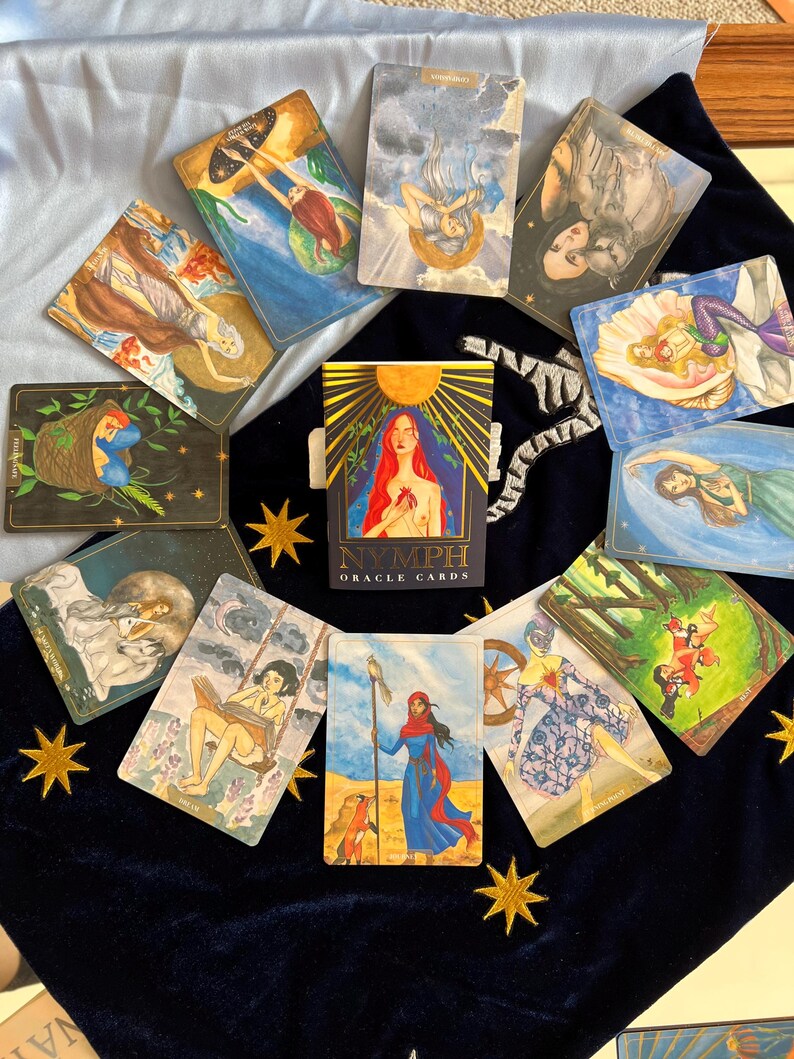 Puede incluir: Un despliegue de cartas de or&aacute;culo con un esquema de color azul, rojo y dorado. Las cartas presentan ilustraciones de mujeres, animales y escenas de la naturaleza. Las cartas est&aacute;n dispuestas en c&iacute;rculo sobre una superficie de terciopelo azul con estrellas doradas. La carta del centro dice "Nymph Oracle Cards".