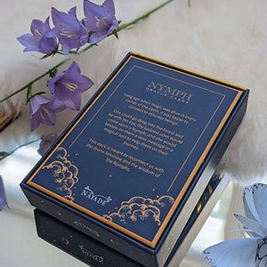 Puede incluir: Una baraja de cartas de or&aacute;culo llamada "Nymph Oracle Cards" con un dise&ntilde;o azul y dorado. La caja presenta una cita sobre la magia que vive en cada rinc&oacute;n de la tierra, y la sabidur&iacute;a de la Ninfa.