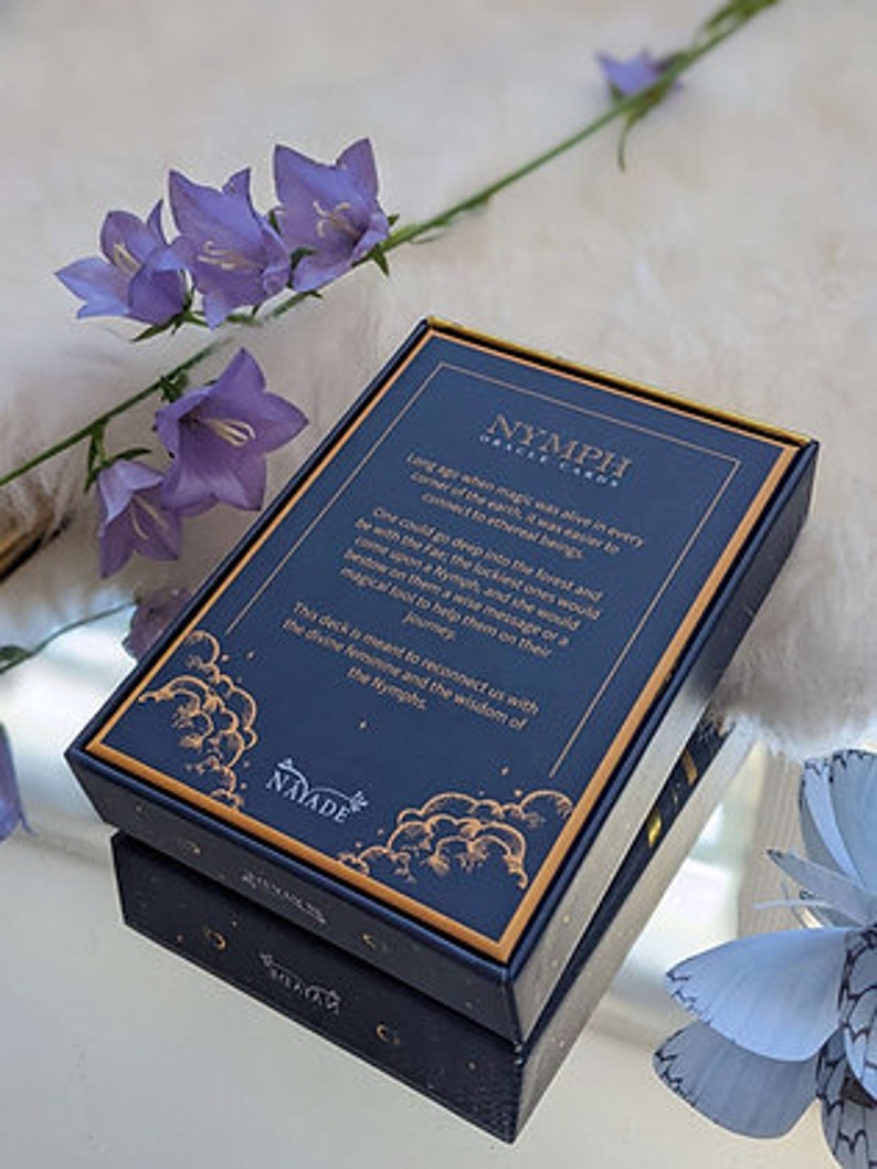 Puede incluir: Una baraja de cartas de or&aacute;culo llamada "Nymph Oracle Cards" con un dise&ntilde;o azul y dorado. La caja presenta una cita sobre la magia que vive en cada rinc&oacute;n de la tierra, y la sabidur&iacute;a de la Ninfa.