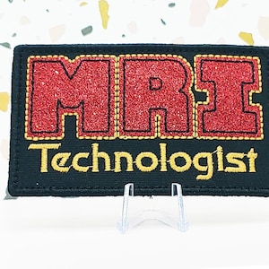 May include: Black rectangular patch with the text "MRI Technologist" in gold and red glitter lettering. The patch is displayed on a white surface with a colorful speckled background.