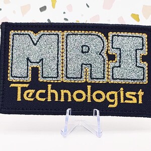 May include: A black embroidered patch with the text "MRI Technologist" in gold thread. The letters "MRI" are in silver glitter and outlined in gold thread.