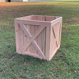 May include: A square wooden planter box with a natural wood finish. The sides feature an X-shaped design. The planter is sitting on a grassy lawn, suggesting outdoor use. The box is approximately 24 inches wide and 24 inches tall.