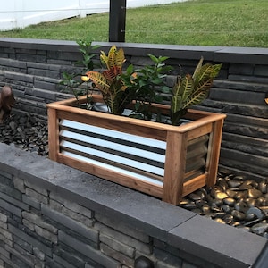 May include: A stone wall with two wooden planter boxes, each with a different type of plant. The planter boxes have a metal liner and are sitting on a bed of black river rocks. There are small lights on the wall.