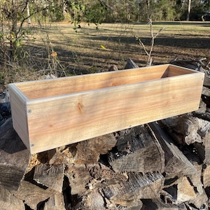 Puede incluir: Una jardinera de madera natural con un tinte marrón claro. La jardinera está sentada sobre una pila de leña apilada.