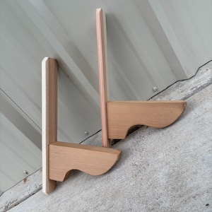 May include: Two unfinished wooden wall shelves. Each shelf has a light-colored vertical support and a horizontal shelf with a curved front edge. The shelves are made of natural wood and are mounted on a gray surface.