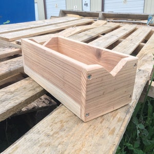 May include: A small wooden box with a V-shaped cutout on the front. The box is made of light-colored wood and has a natural finish. It is sitting on a wooden pallet.