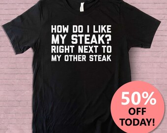 Könnte beinhalten: Schwarzes T-Shirt mit weißem Text, der lautet: "Wie mag ich mein Steak? Direkt neben meinem anderen Steak." Das T-Shirt befindet sich auf einem rosafarbenen Ziegelhintergrund mit einem roten Kreis, auf dem "50% Rabatt heute!" steht. Der Text "Cosmic Rebel" befindet sich in der oberen rechten Ecke des Bildes.