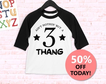 Puede incluir: Una camiseta de béisbol blanca y negra con el texto "Ain't nothin' but a 3 thang" en negro. La camiseta está colgada de una percha negra contra una pared de ladrillo blanco. Un círculo rosa con el texto "50% de descuento hoy!" está en la esquina inferior derecha de la imagen.