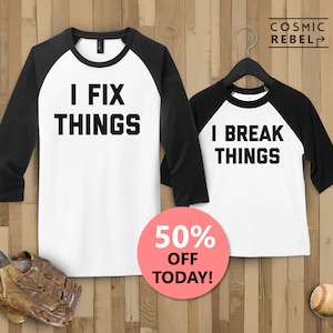 May include: Two baseball-style shirts with black sleeves and white bodies. The larger shirt says "I FIX THINGS" and the smaller shirt says "I BREAK THINGS".