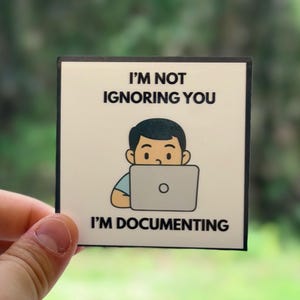Może przedstawiać: Kwadratowa naklejka z czarną obwódką przedstawiająca kreskówkową ilustrację osoby za laptopem. Tekst brzmi: "I'M NOT IGNORING YOU I'M DOCUMENTING". Tło jest rozmyte zielone.