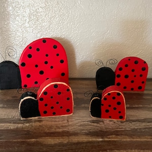 May include: Five wooden ladybug ornaments, painted red with black dots and black wings. The ornaments are arranged in a row on a wooden surface.
