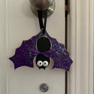 May include: A Halloween-themed bat ornament with purple glitter wings and a black body. The bat has white eyes and a purple ribbon bow. It hangs from a black ribbon on a white door.