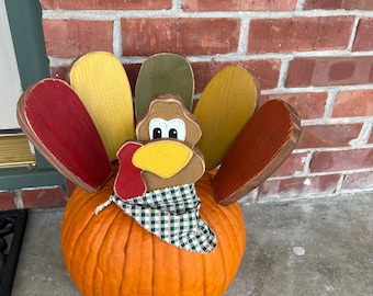 May include: A Thanksgiving decoration featuring a carved wooden turkey head and colorful tail feathers attached to an orange pumpkin. The turkey wears a green and white checkered scarf. The background is a red brick wall.