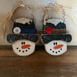 May include: Two wooden snowman ornaments. Each snowman has a white face with black dot eyes and a carrot nose. One snowman has a blue hat with a white button, the other has a plaid hat with a red button. Both have glitter and straw hangers.