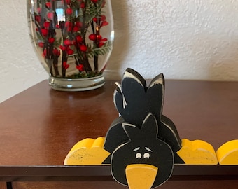 May include: A black crow stocking holder with yellow wings and beak. The crow is peeking over the edge of a wooden surface.