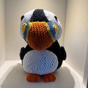 May include: A handmade crochet puffin bird with a white, black, orange, yellow, and blue color scheme. The puffin has a large orange beak, black eyes, and small orange feet. The body is primarily white and black.