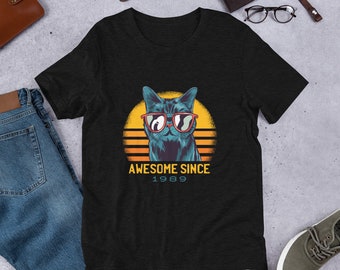 May include: Black t-shirt featuring a blue cat wearing red sunglasses, set against a yellow and orange sunset design. The text "AWESOME SINCE 1989" is printed below the cat graphic. The shirt is made of a soft material.