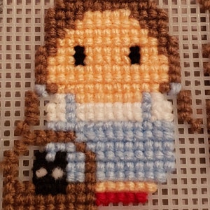 May include: A cross-stitch pattern of a girl with brown hair wearing a blue dress and holding a brown and black camera.