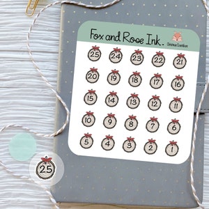 May include: A sheet of 25 Christmas countdown stickers featuring a woodgrain circle design with a red bow and a white number. The stickers are arranged in a grid pattern and are labeled with numbers 1 through 25.