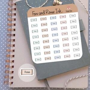 May include: A sheet of stickers with the text "Fox and Rose Ink. Protein Bars" at the top. The stickers are in the shape of a rectangle and have the word "Protein" written on them. The stickers are in three different colors: light blue, light green, and light pink.