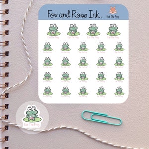 May include: A sheet of stickers featuring a cartoon frog sitting on a lily pad. The frog is green with pink accents and the text "Eat The Frog" is written below each frog. The sheet is white with a light blue border and the text "Fox and Rose Ink." is written at the top.