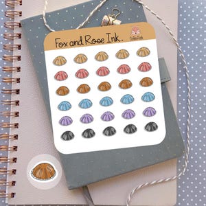 May include: A sheet of colorful stickers featuring a seashell design in various colors, including tan, pink, brown, blue, purple, and gray. The stickers are arranged on a white background with the text "Fox and Rose Ink." and "Coffee Pods."