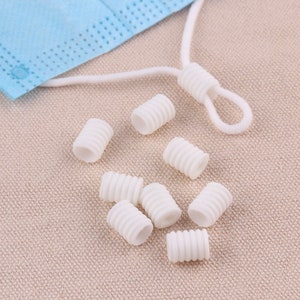 May include: A collection of white plastic cord adjusters with a ribbed design, alongside a blue face mask and white elastic cord. These small cylindrical pieces are designed to adjust the fit of face masks.