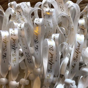 May include: A pile of white satin ribbon wristbands with gold lettering. The text on the wristbands reads "The Beckford" and "June 7, 2025".