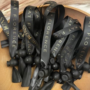 May include: A close-up of a pile of black satin ribbon with gold lettering that reads "KAT FLORENCE". The ribbon is tied with black rubber stoppers.