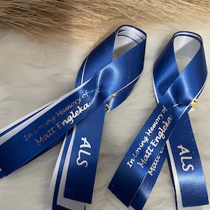 May include: Two blue and white ribbon pins with gold lettering. The ribbons say "In loving memory of Matt Engleka" and "ALS".