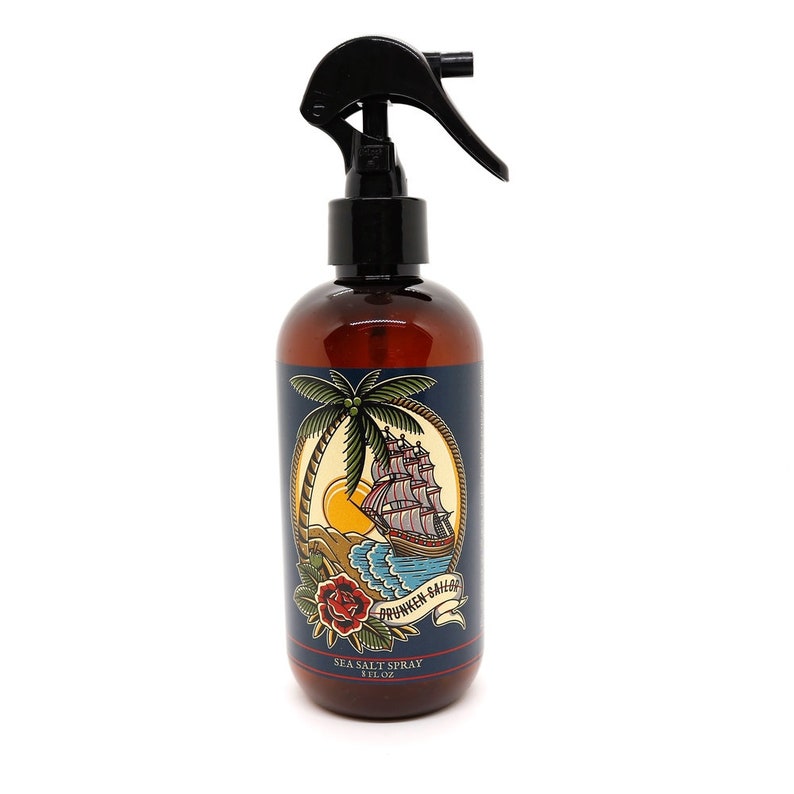 May include: A brown bottle with a black spray nozzle and a blue label. The label features a nautical themed illustration of a ship sailing past a palm tree and a red rose. The text on the label reads "Drunken Sailor Sea Salt Spray 8 fl oz".