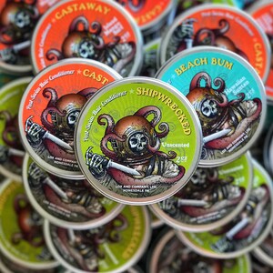 May include: A close-up of several round, metal tins with colorful labels. The labels feature illustrations of an octopus and a diver wearing a helmet. The text on the labels includes "Shipwrecked", "Unscented", "2 oz", "Beach Bum", "Hawaiian Suntan", "Castaway", and "Post-Swim Hair Conditioner".