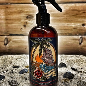 May include: A brown glass bottle with a black spray nozzle and a label featuring a nautical scene with a ship, palm tree, and the text "Drunken Sailor Sea Salt Spray 8 fl oz". The bottle is surrounded by gold coins on a sandy surface.