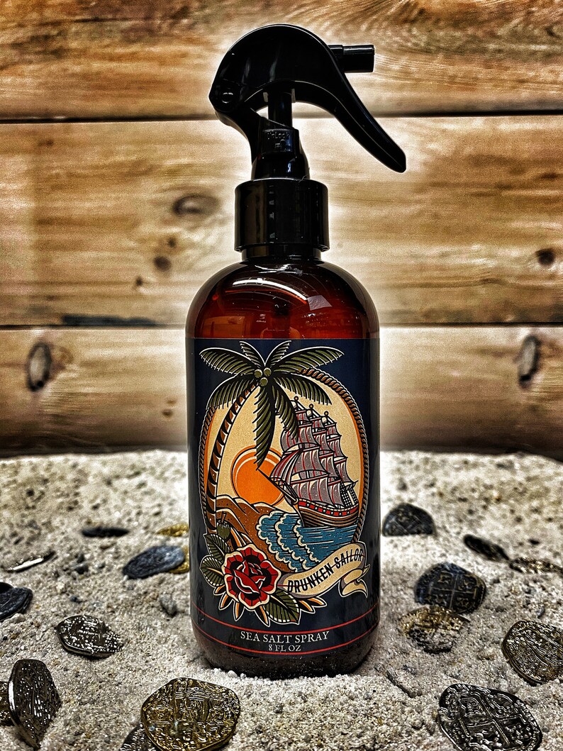 May include: A brown glass bottle with a black spray nozzle and a label featuring a nautical scene with a ship, palm tree, and the text "Drunken Sailor Sea Salt Spray 8 fl oz". The bottle is surrounded by gold coins on a sandy surface.