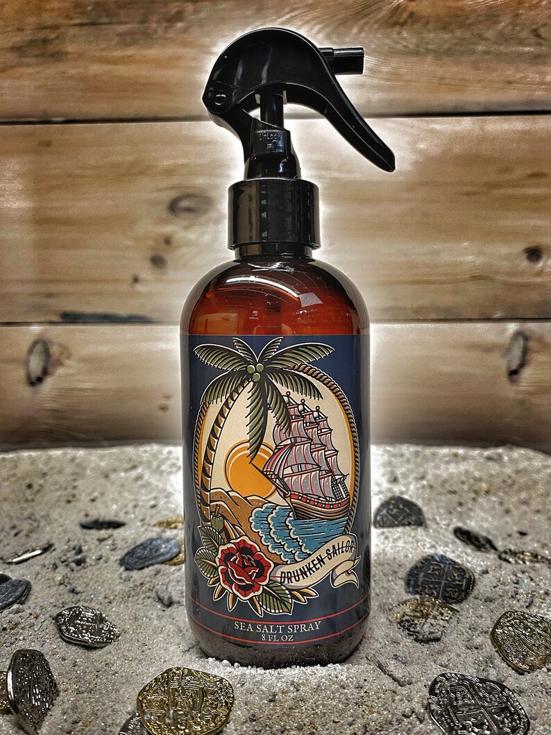 May include: A brown glass bottle with a black spray nozzle. The bottle has a label with a nautical theme, featuring a ship, palm tree, and the text "Drunken Sailor Sea Salt Spray 8 fl oz".