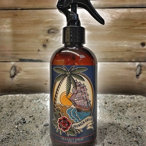 May include: A brown glass bottle with a black spray nozzle and a blue label. The label features a nautical themed illustration of a ship, palm tree, and a rose. The text on the label reads "Drunken Sailor Sea Salt Spray 8 fl oz".
