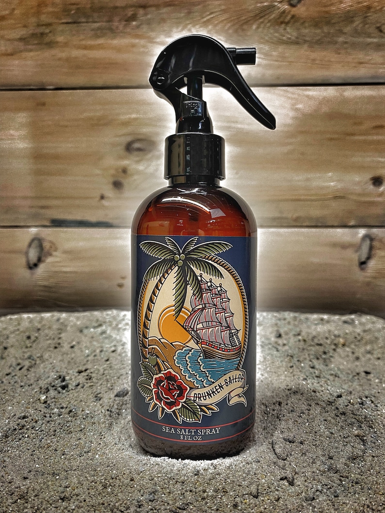 May include: A brown glass bottle with a black spray nozzle and a blue label. The label features a nautical themed illustration of a ship, palm tree, and a rose. The text on the label reads "Drunken Sailor Sea Salt Spray 8 fl oz".