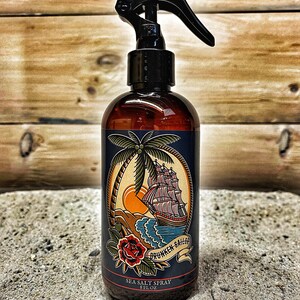 May include: A brown glass bottle with a black spray nozzle and a blue label. The label features a nautical illustration of a ship sailing on the ocean with a palm tree and a red rose. The text on the label reads "Drunken Sailor Sea Salt Spray 8 FL OZ".